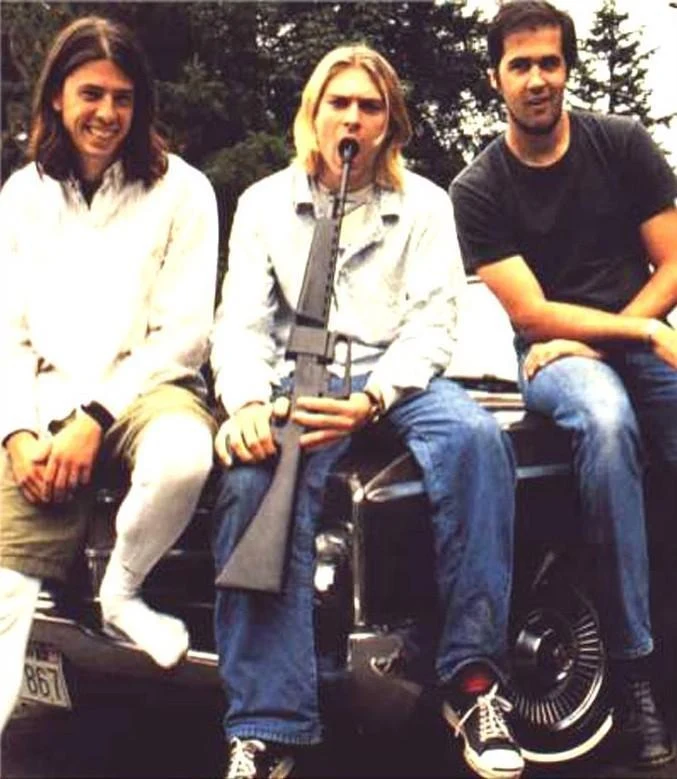 Kurt Cobain, Dave Grohl y Krist Novoselic sentados sobre el capó de un auto, Cobain sosteniendo un rifle